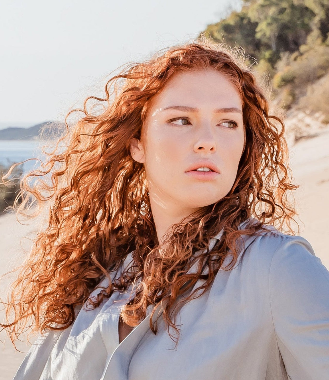 A woman with long curly red hair sits barefoot on sand dunes next to a large piece of driftwood, wearing a light gray long-sleeve robe. The background features a calm blue ocean, distant islands, and green forested hills under a clear sky.