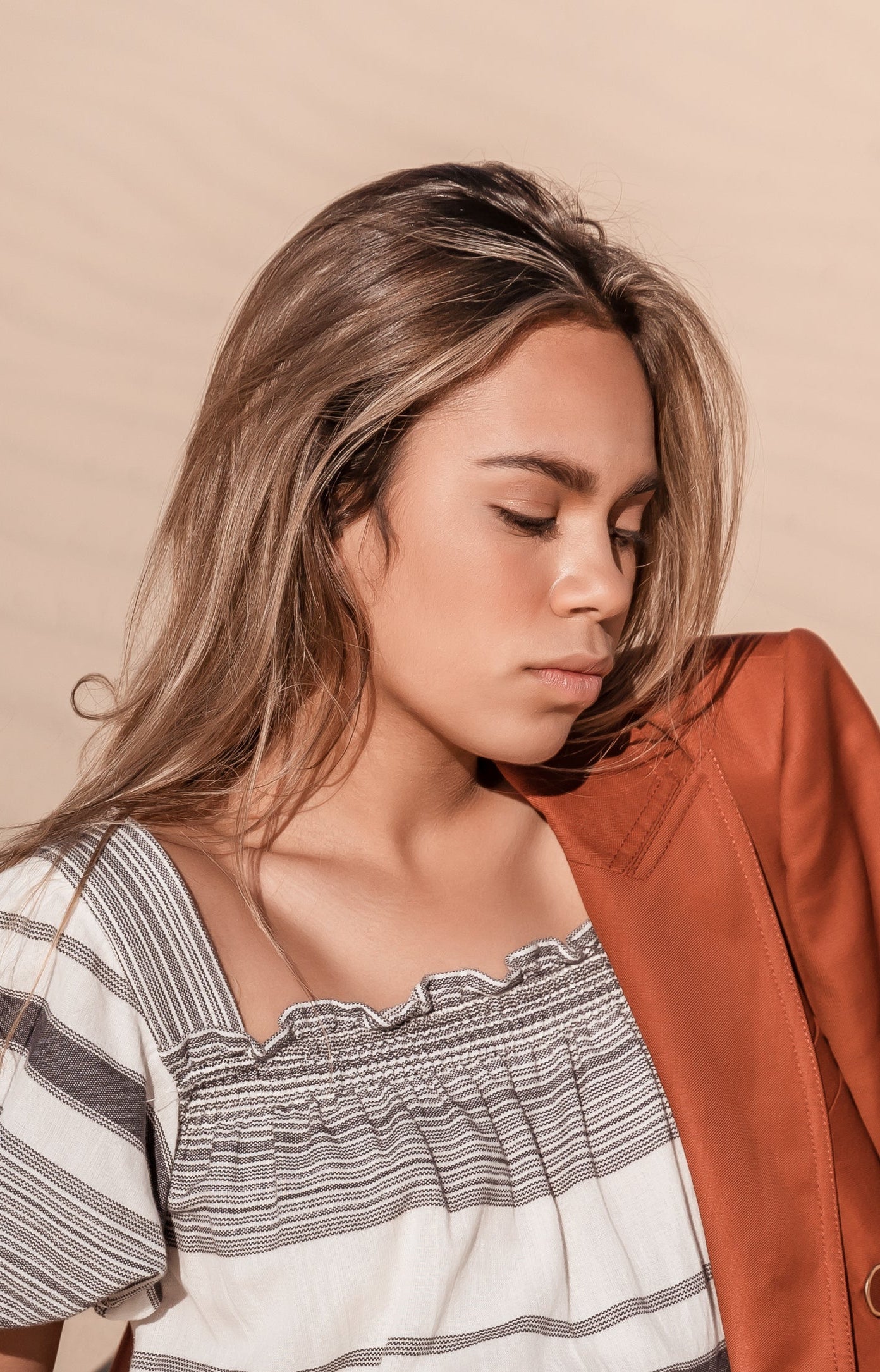A woman sitting in a sandy desert setting, wearing a white smocked dress with gray stripes and a rust-orange satin blazer draped over her shoulder.